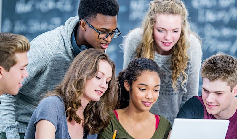 Group of six diverse students collaborating around a laptop in a classroom, discussing work and looking engaged in a shared learning activity.