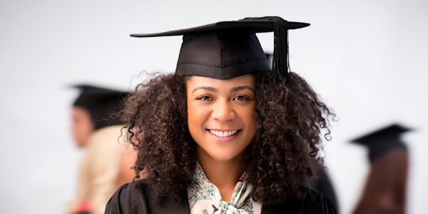 Smiling graduate wearing a mortarboard and gown, with other graduates blurred in the background.