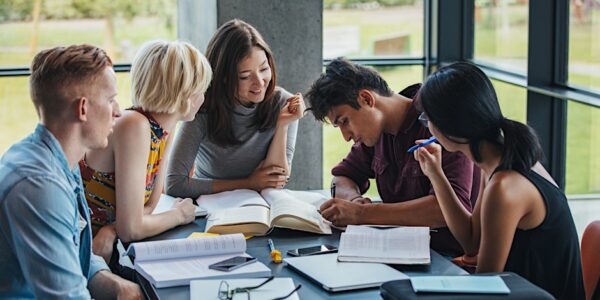 Group of students sitting around a table studying together, sharing books and notes in a bright indoor space.