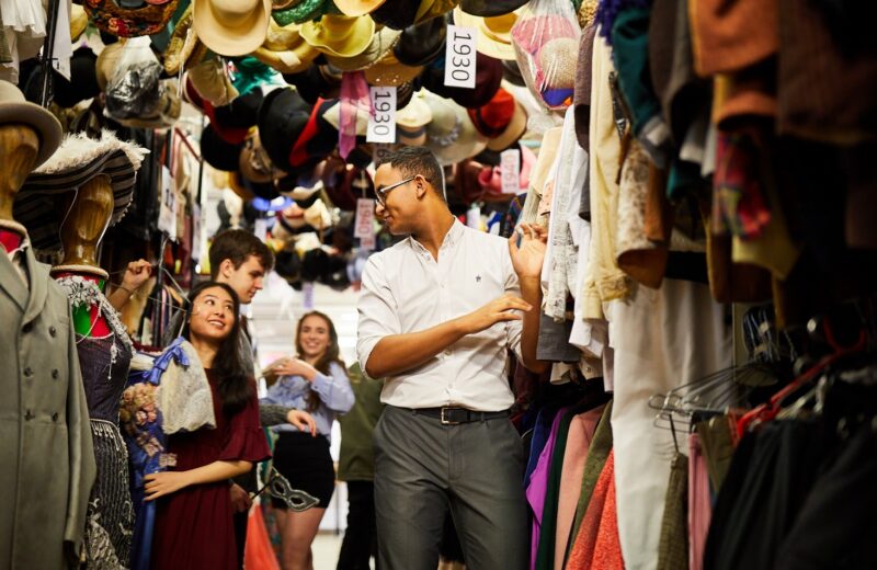 People browsing clothing on rails in a busy indoor market, with colourful garments and hats displayed closely together.